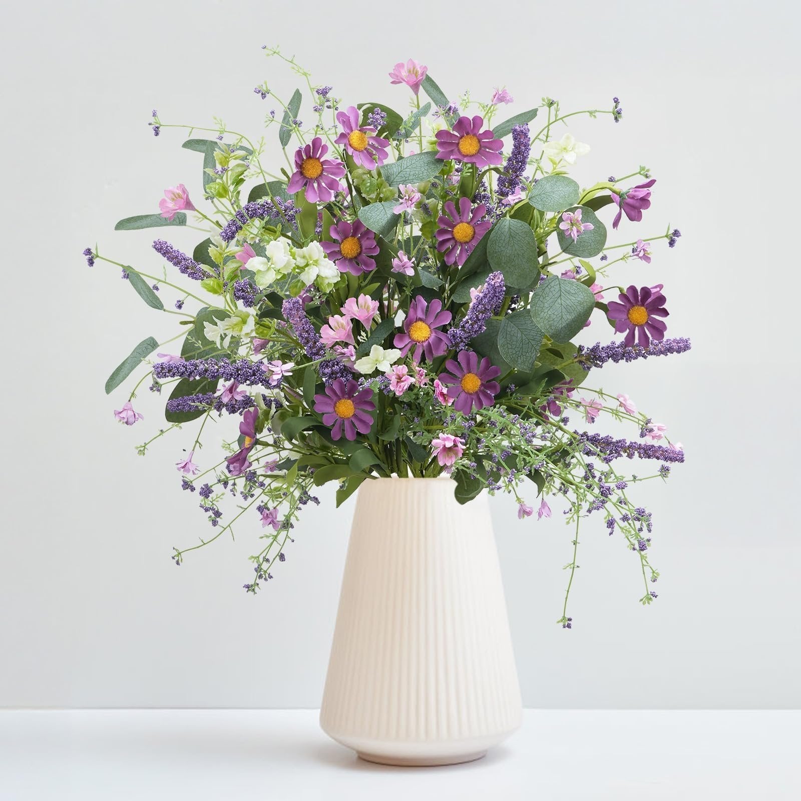 Artificial spring flower bouquet with purple daisies, lavender, and eucalyptus leaves in a cream vase.
