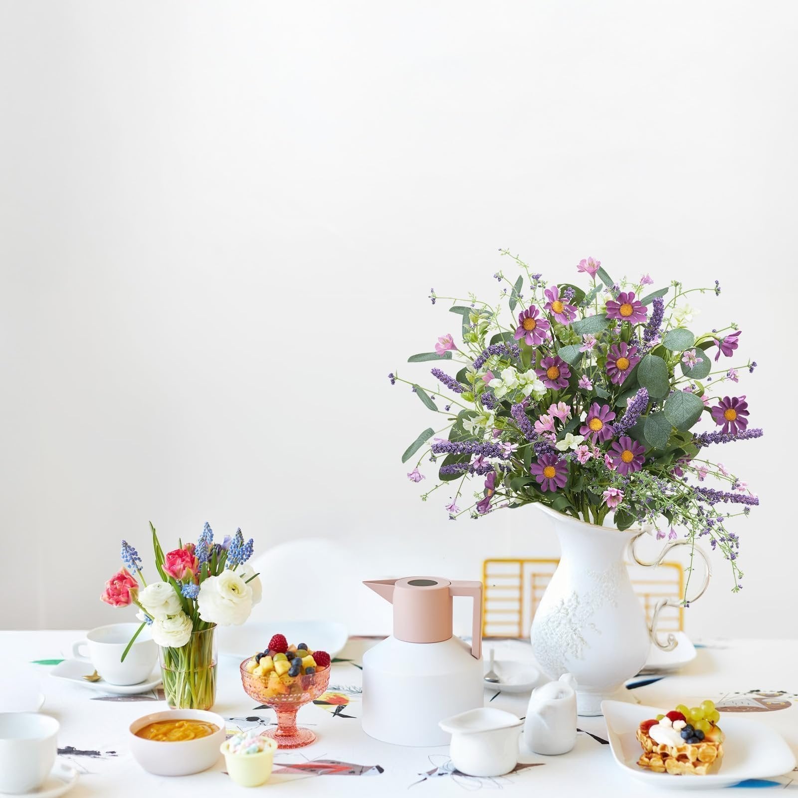 Artificial spring flower bouquet with purple daisies and lavender in a white pitcher on a table with breakfast items