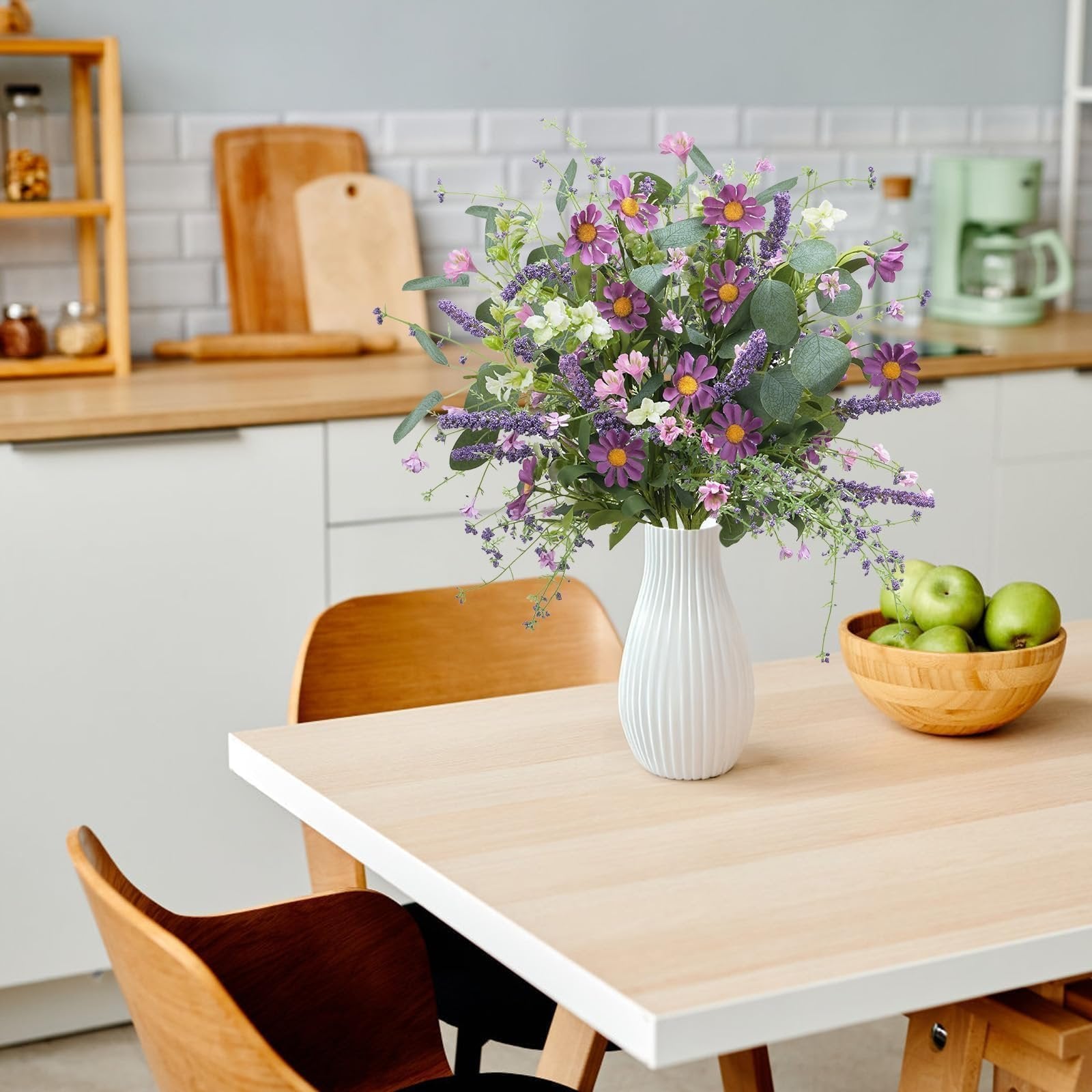 Artificial spring flower bouquet with purple daisies and lavender in a white vase on a table with green apples.
