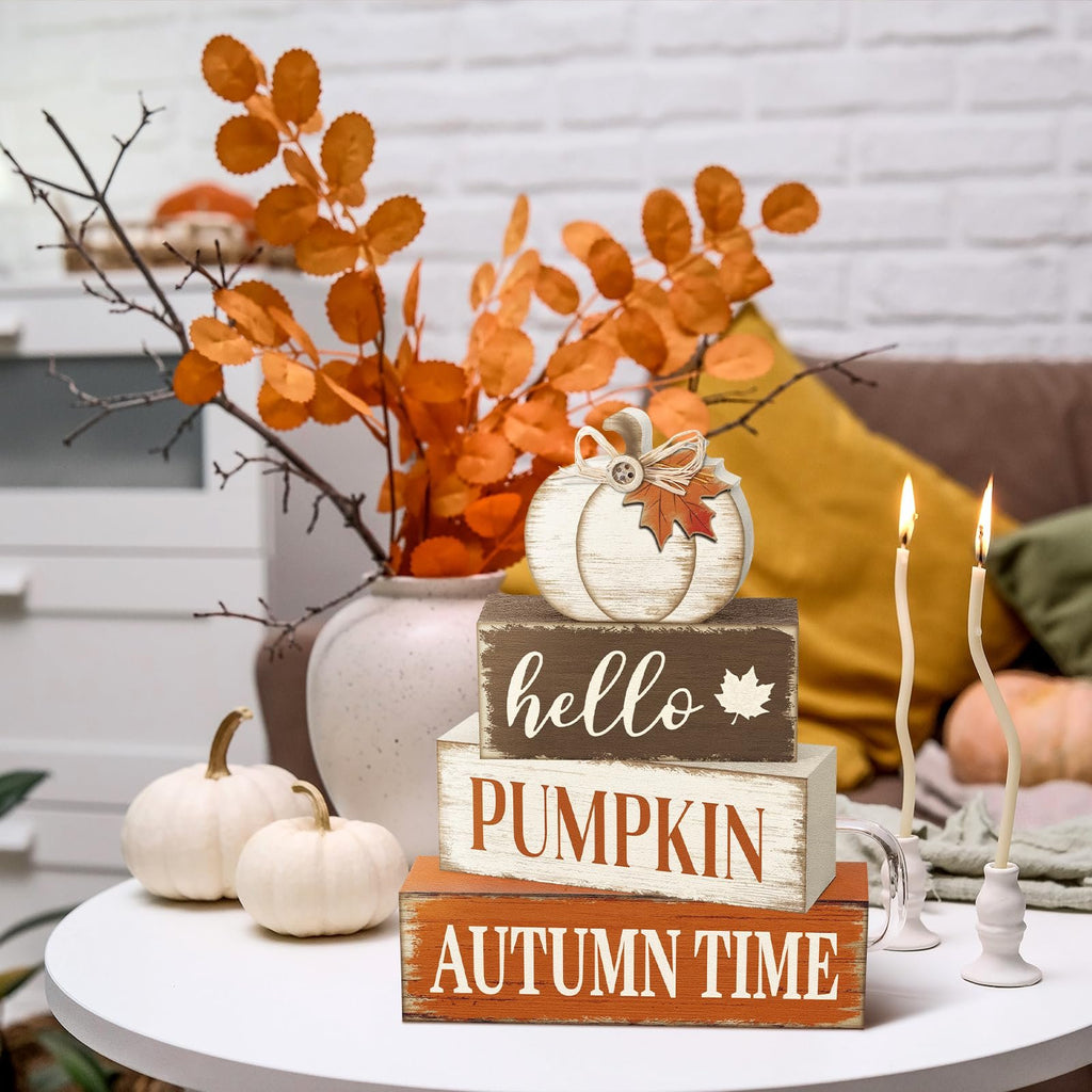 Stack of three wooden signs with autumn phrases, topped with a decorative pumpkin, on a table with white pumpkins and candles.