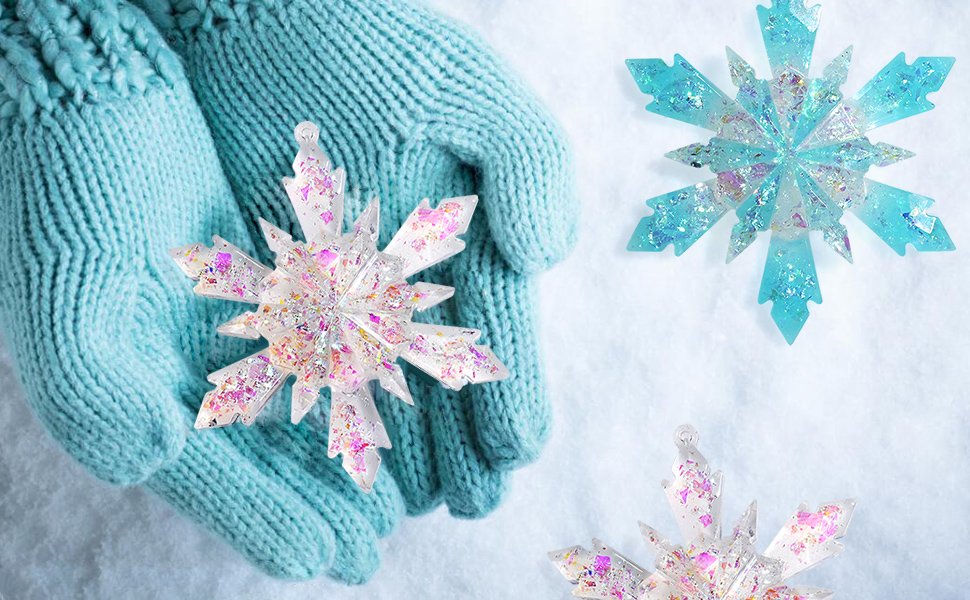 Hands wearing blue knitted gloves hold a clear snowflake ornament with pink and purple glitter, surrounded by snow and other snowflake ornaments.