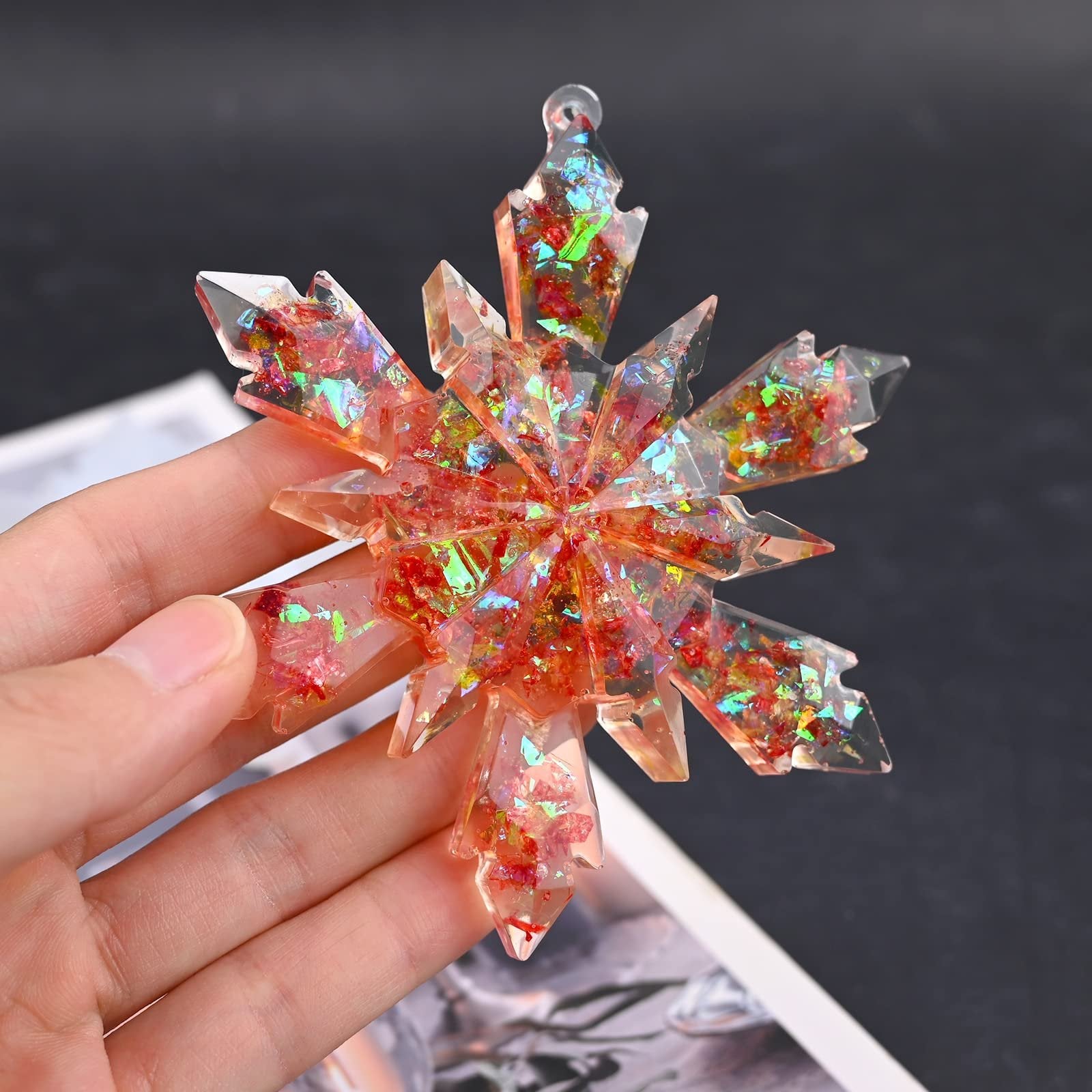 Hand holding a clear snowflake-shaped resin ornament with red and iridescent glitter inclusions.