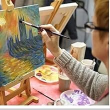 Person painting a colorful landscape on canvas with a brush, easel, and paint supplies visible