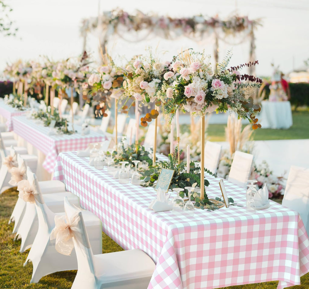 Pink gingham disposable plastic tablecloth set on a long table decorated for an outdoor event with floral centerpieces and white chairs.