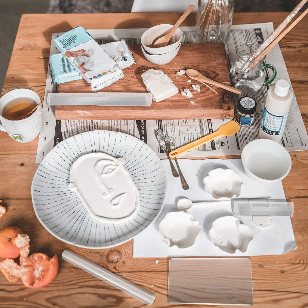 Clay sculpting setup with face impression on plate, clay blocks, roller, and tools on newspaper