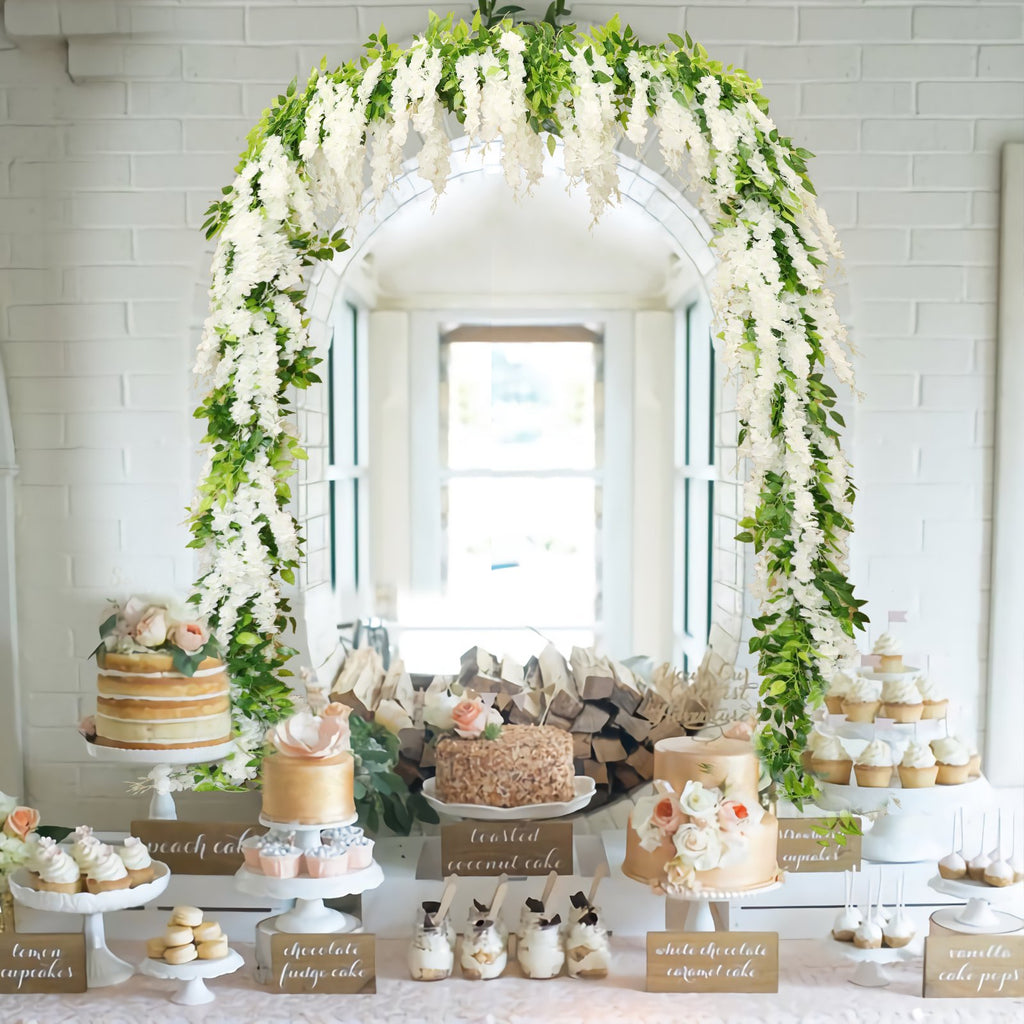 White wisteria garland draped over an archway above a dessert table with cakes and cupcakes.