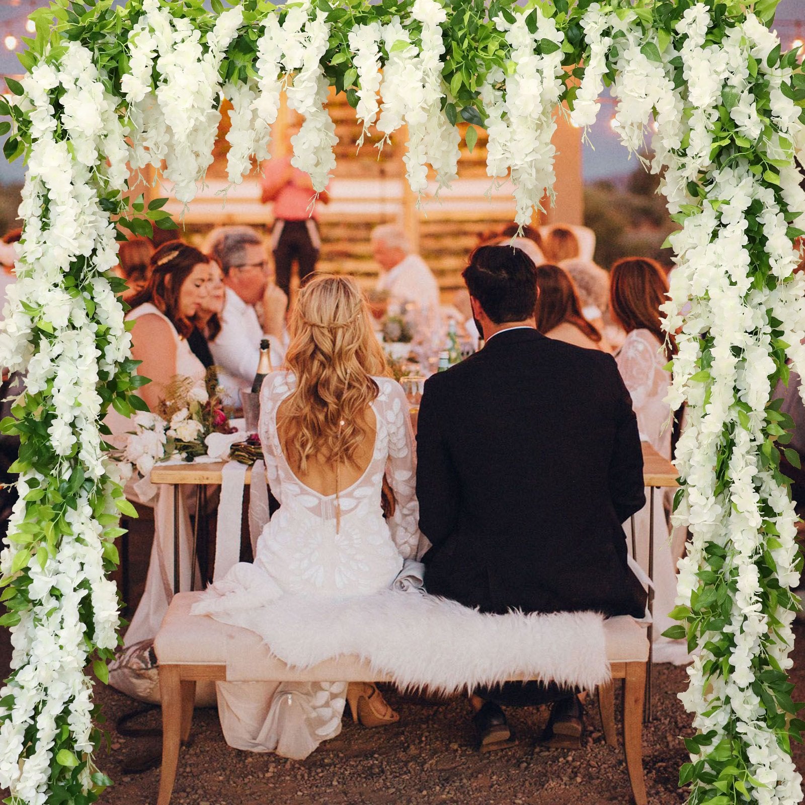 Wedding reception scene with a bride and groom seated at a table, framed by white wisteria flower garlands.