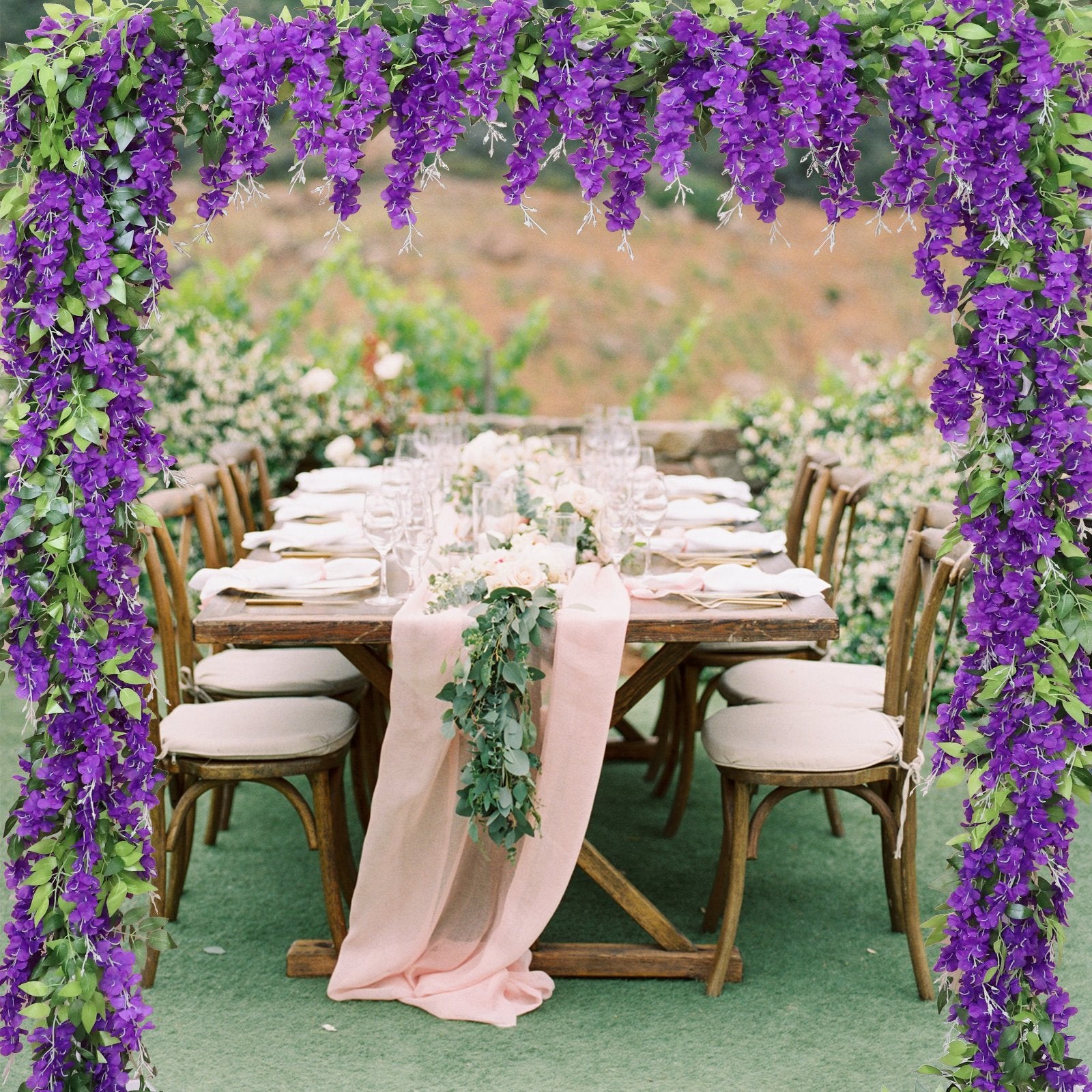 Purple wisteria garlands framing an outdoor wedding reception table setting with floral centerpieces and place settings.