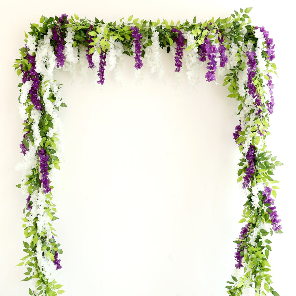 White and purple wisteria garlands draped over a white wall, forming an arch shape.