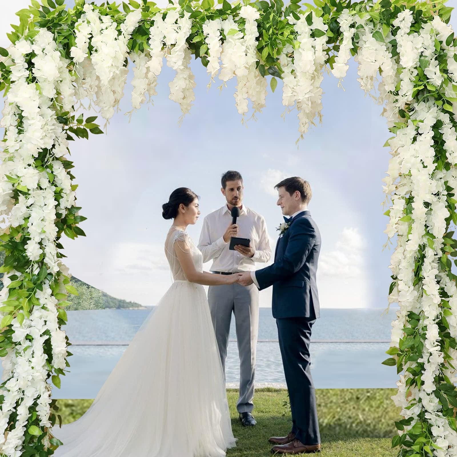 Wedding ceremony with a bride and groom holding hands under a white wisteria flower arch with green leaves, overlooking the ocean.