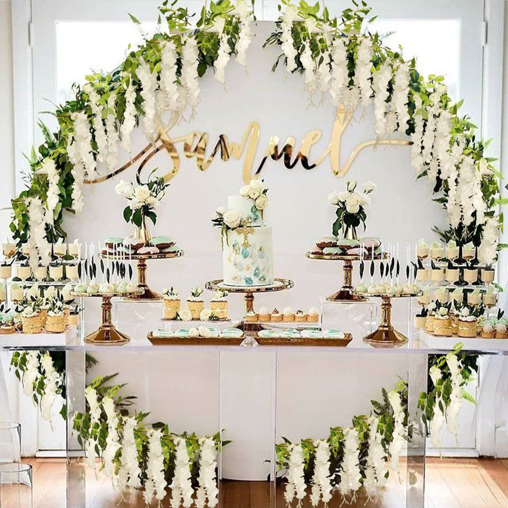 White wisteria garlands draped over a dessert table with a cake and cupcakes, with a gold name sign in the background.