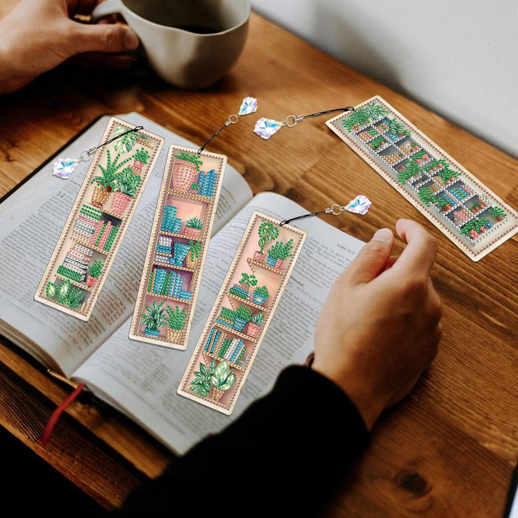 Four diamond painting bookmarks with plant and bookshelf designs resting on an open book.