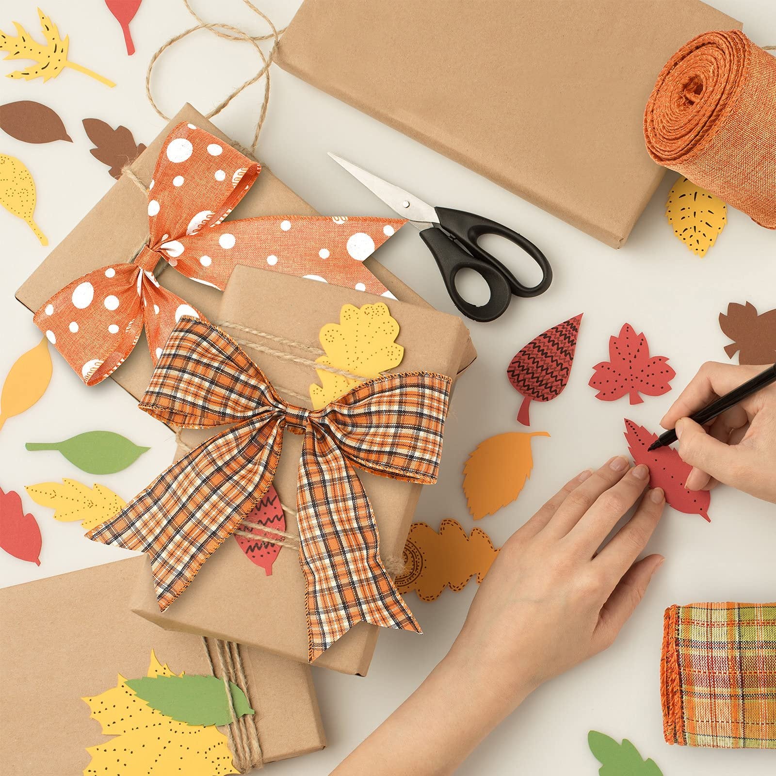 Hands decorating gifts with plaid ribbon and fall leaf cutouts, scissors and ribbon roll nearby