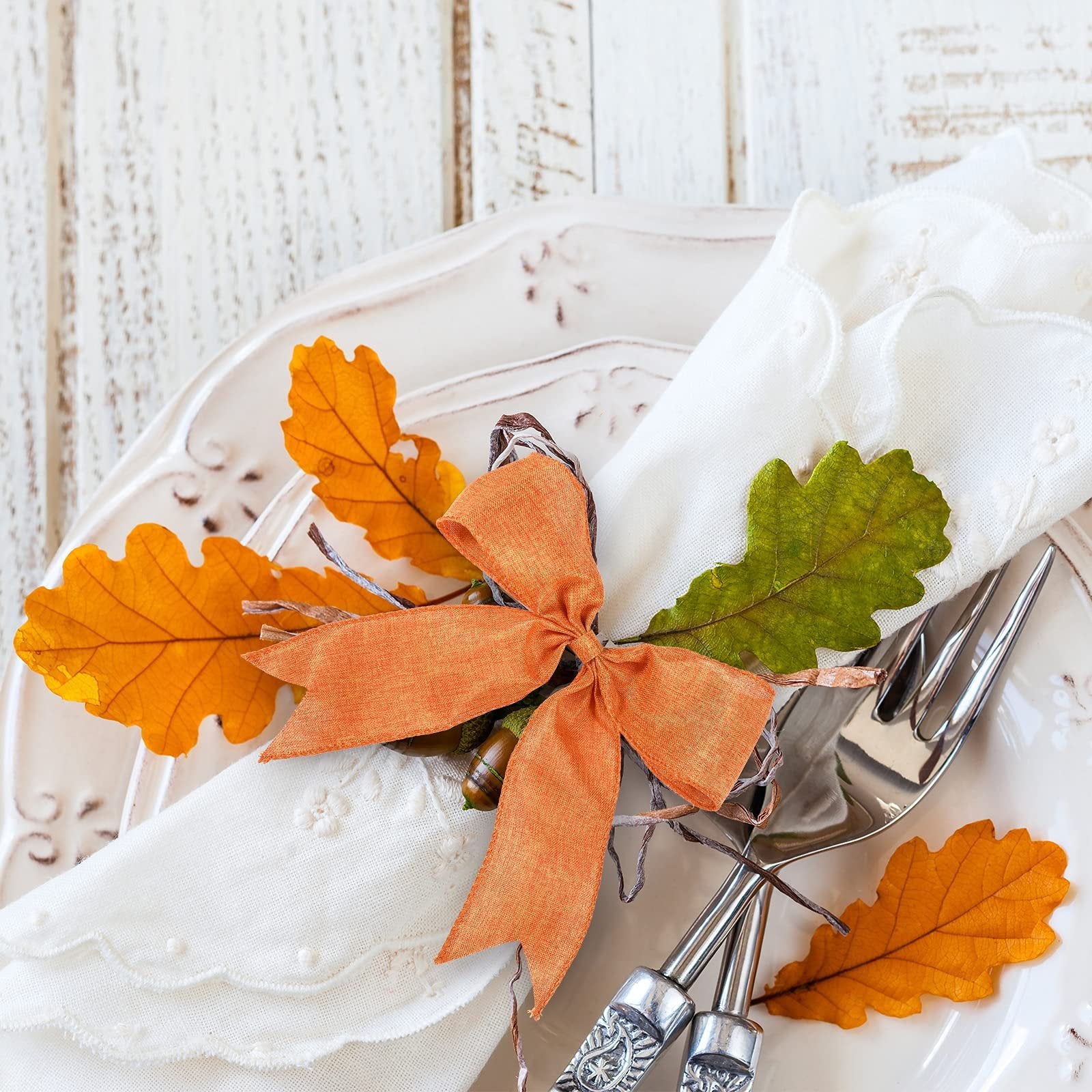 Table setting with a white napkin adorned with an orange bow, acorns, and fall leaves, next to silverware.