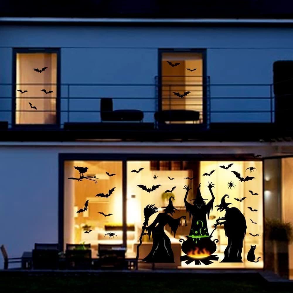 Halloween window clings with witches, bats, spiders, and cauldron silhouettes applied to a house exterior at night.