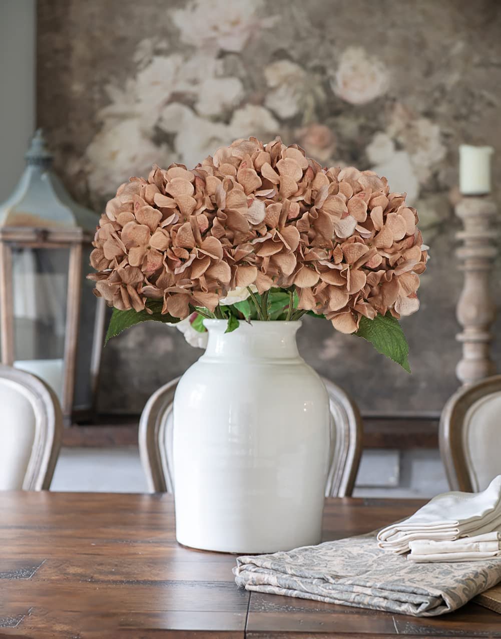Four brown artificial hydrangea flower stems in a white vase on a wooden table.