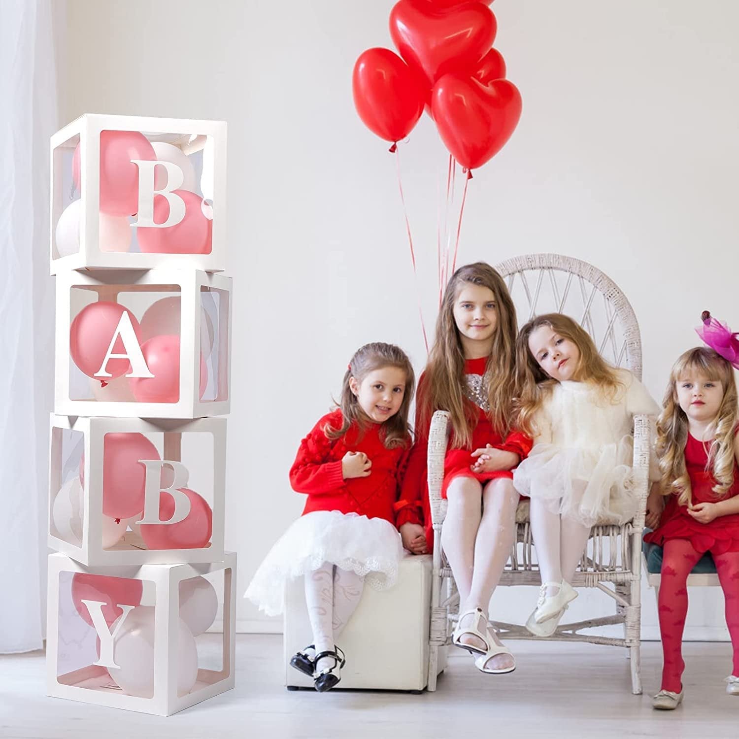Four transparent white balloon boxes stacked with pink and white balloons inside, spelling BABY, with four young girls in red dresses and heart balloons.