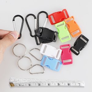 Hand holding carabiner clips next to colorful buckles, D-rings, and a measuring tape on a white background.