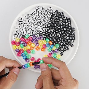 Hands stringing colorful beads and letter beads onto black cord from a white plate, part of a paracord bracelet kit.