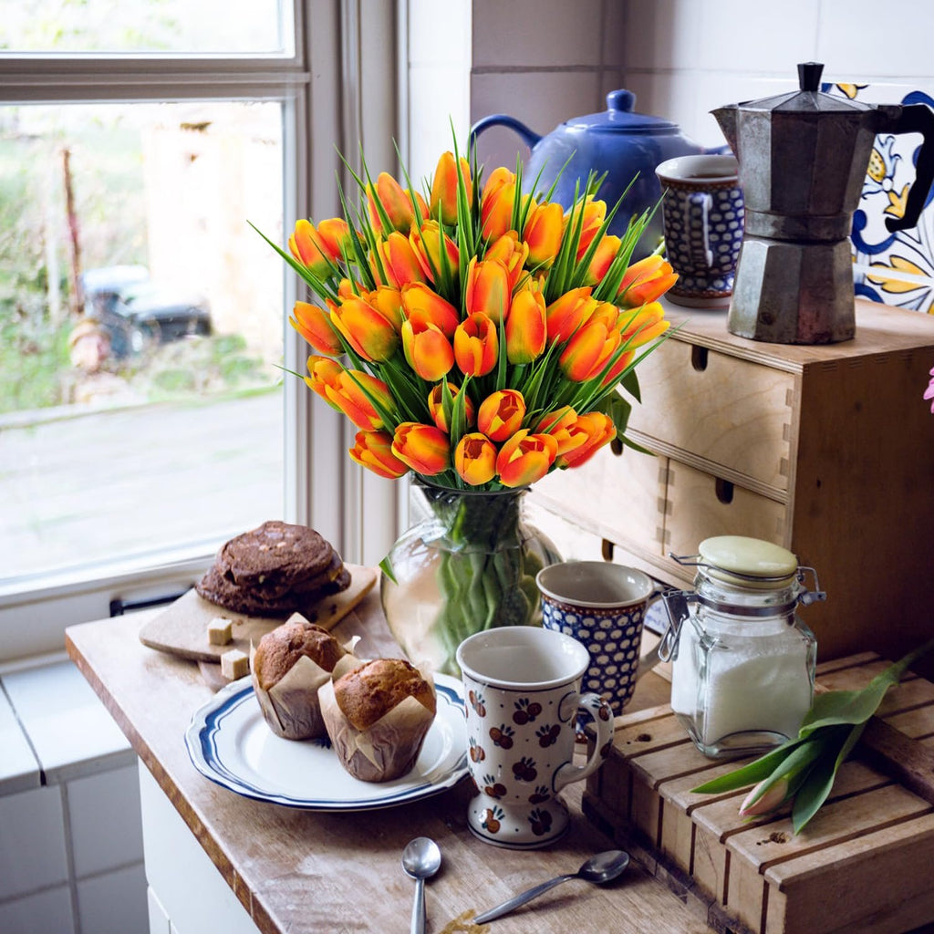 A bouquet of orange artificial tulips in a glass vase on a wooden table with muffins and coffee accessories