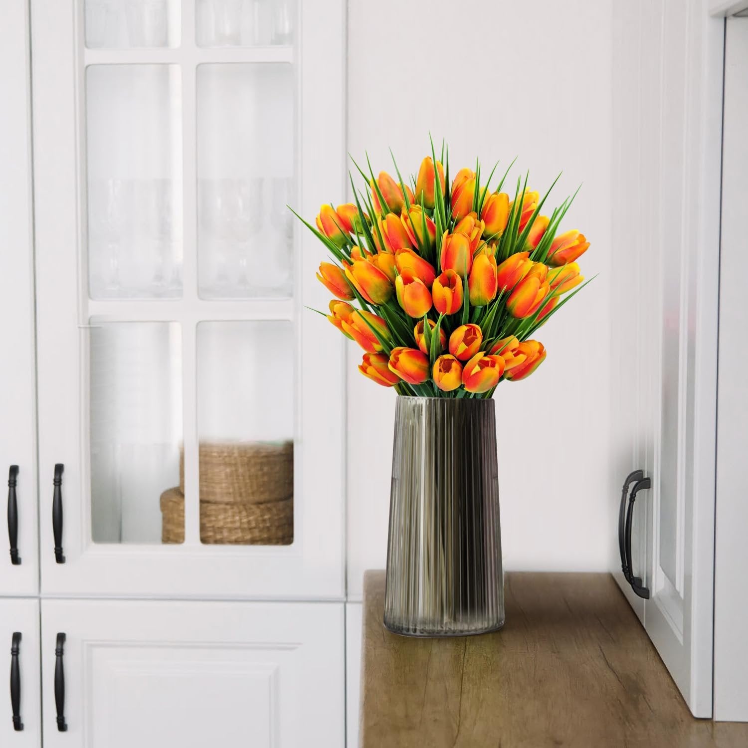 Bundle of 40 orange artificial tulips in a ribbed glass vase on a wooden surface in front of white cabinetry.