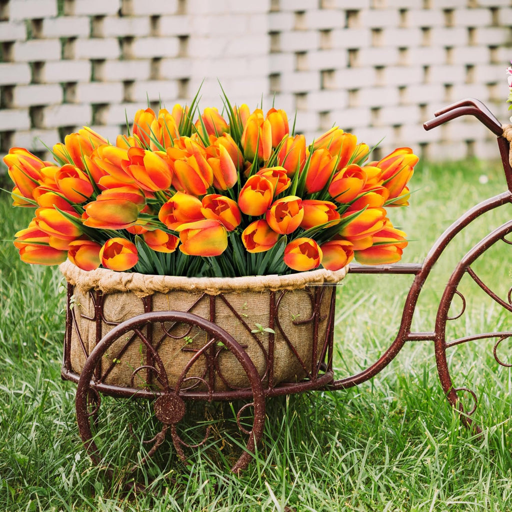 Artificial orange and yellow tulips in a decorative bicycle planter on grass with a brick wall background