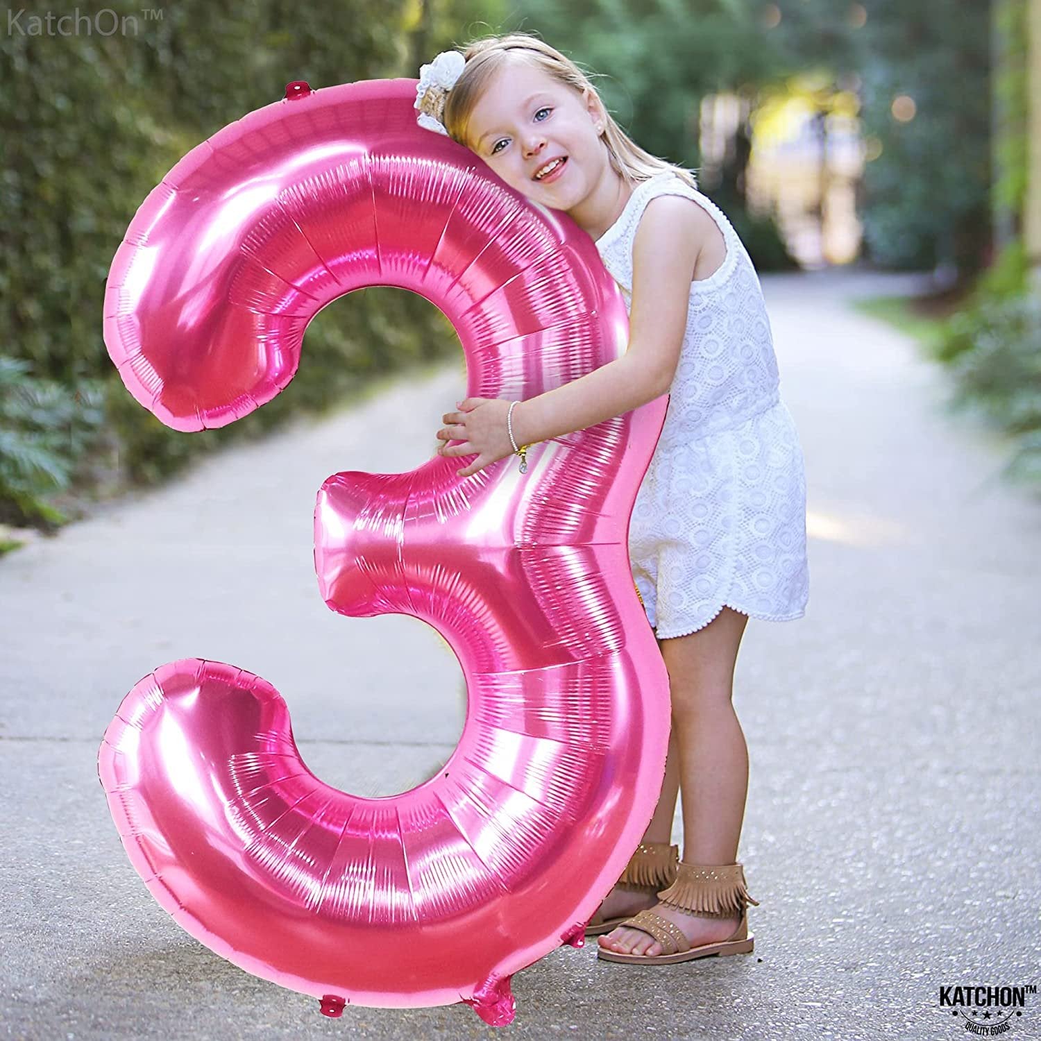 Young girl hugging a large pink number 3 foil balloon outdoors.