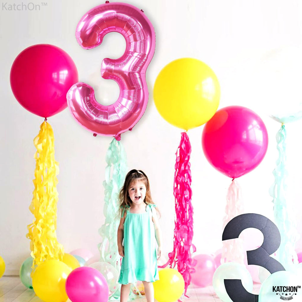 A young girl stands among colorful balloons and streamers, with a large pink number 3 balloon.