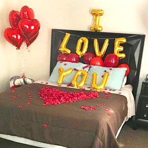 Valentine's Day bedroom decor with heart balloons, 'I LOVE YOU' letter balloons, and silk rose petals arranged on a bed.