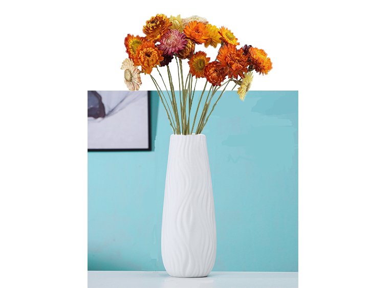 A bouquet of dried daisies in various orange and yellow hues arranged in a white textured vase against a teal background.