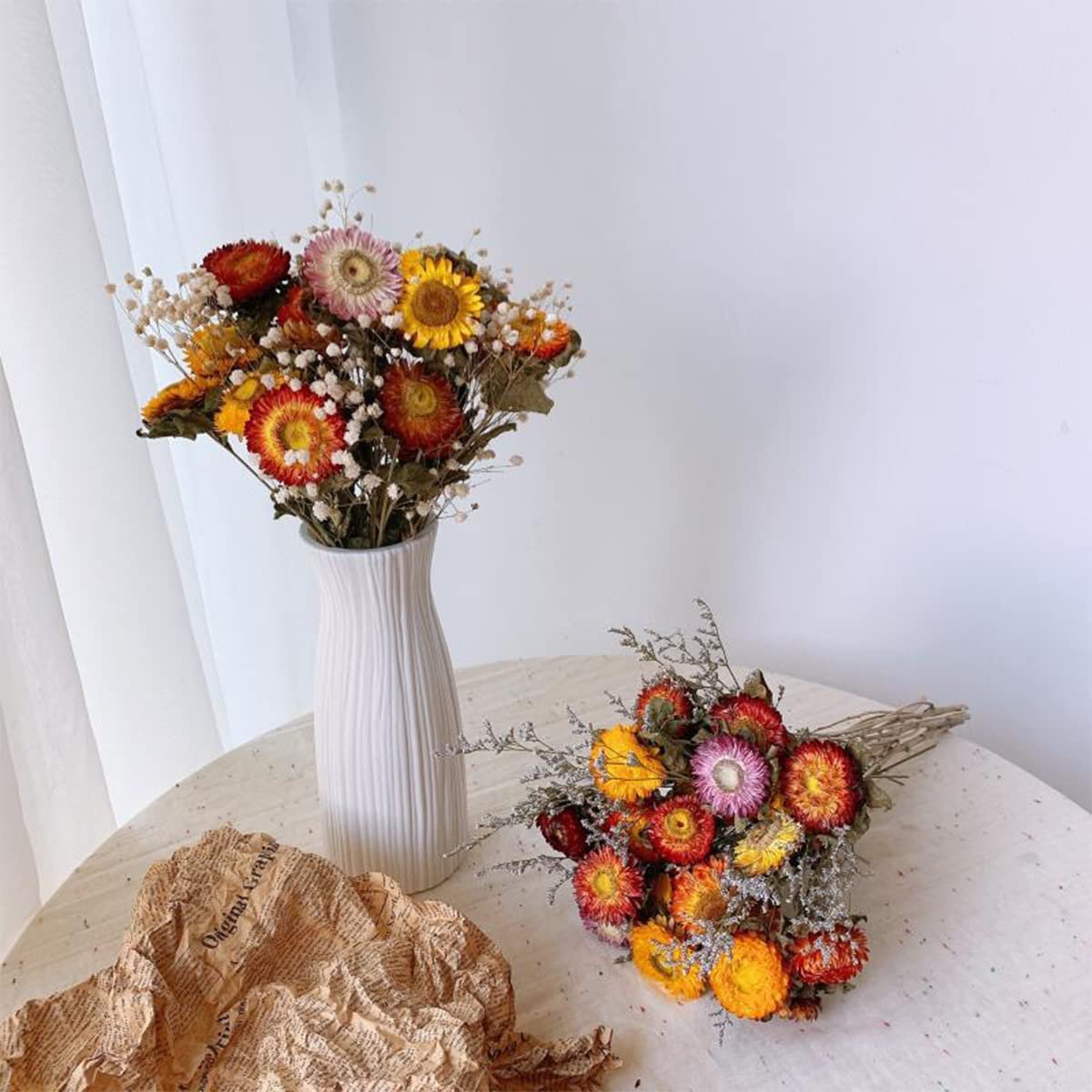 Two bouquets of colorful dried daisies with baby's breath, one in a white vase, the other on a table.