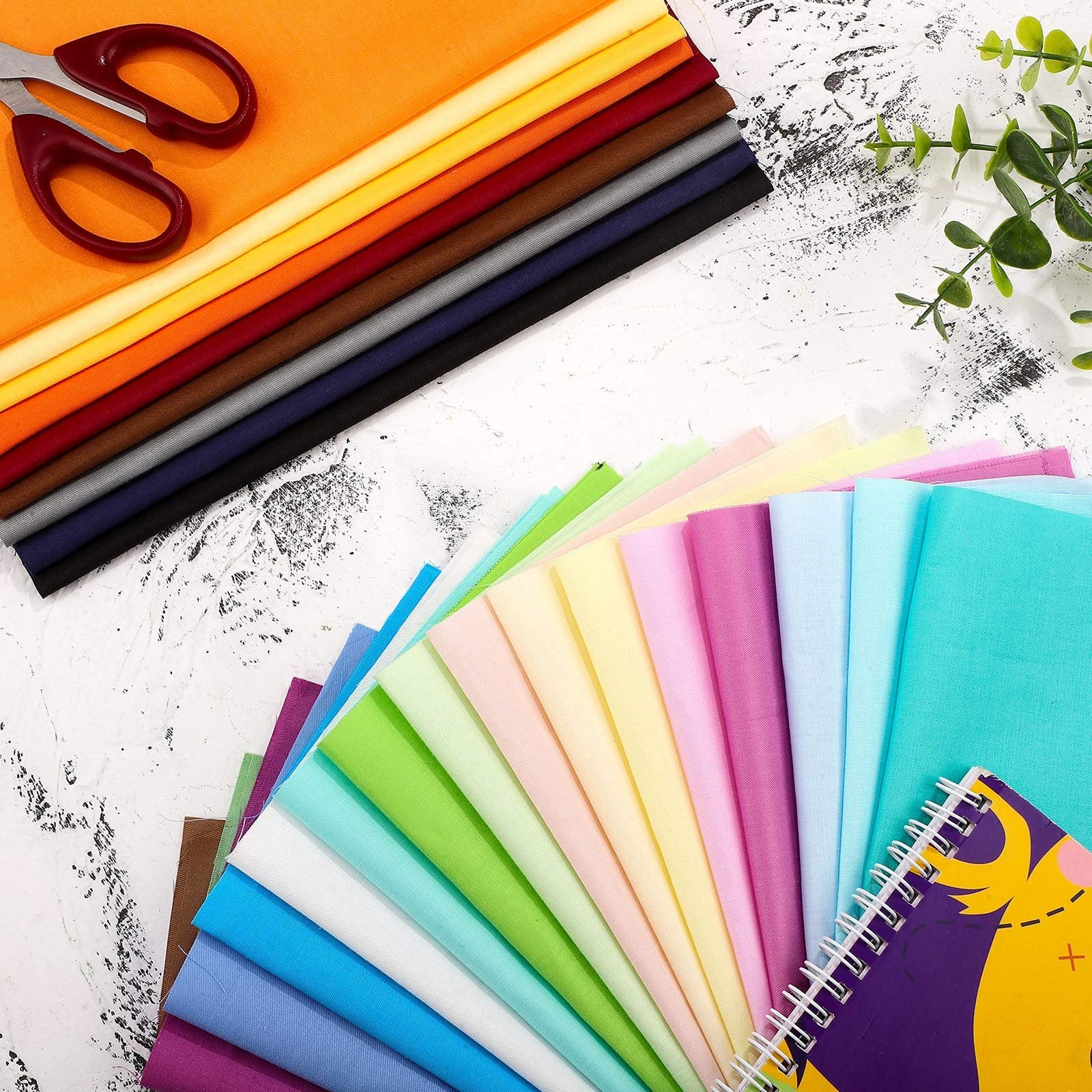 Stack of fabric squares in various colors, with scissors and a notebook, top-down view.