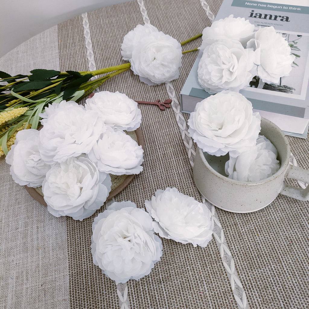 White artificial rose heads scattered on a textured tablecloth with a book and a mug
