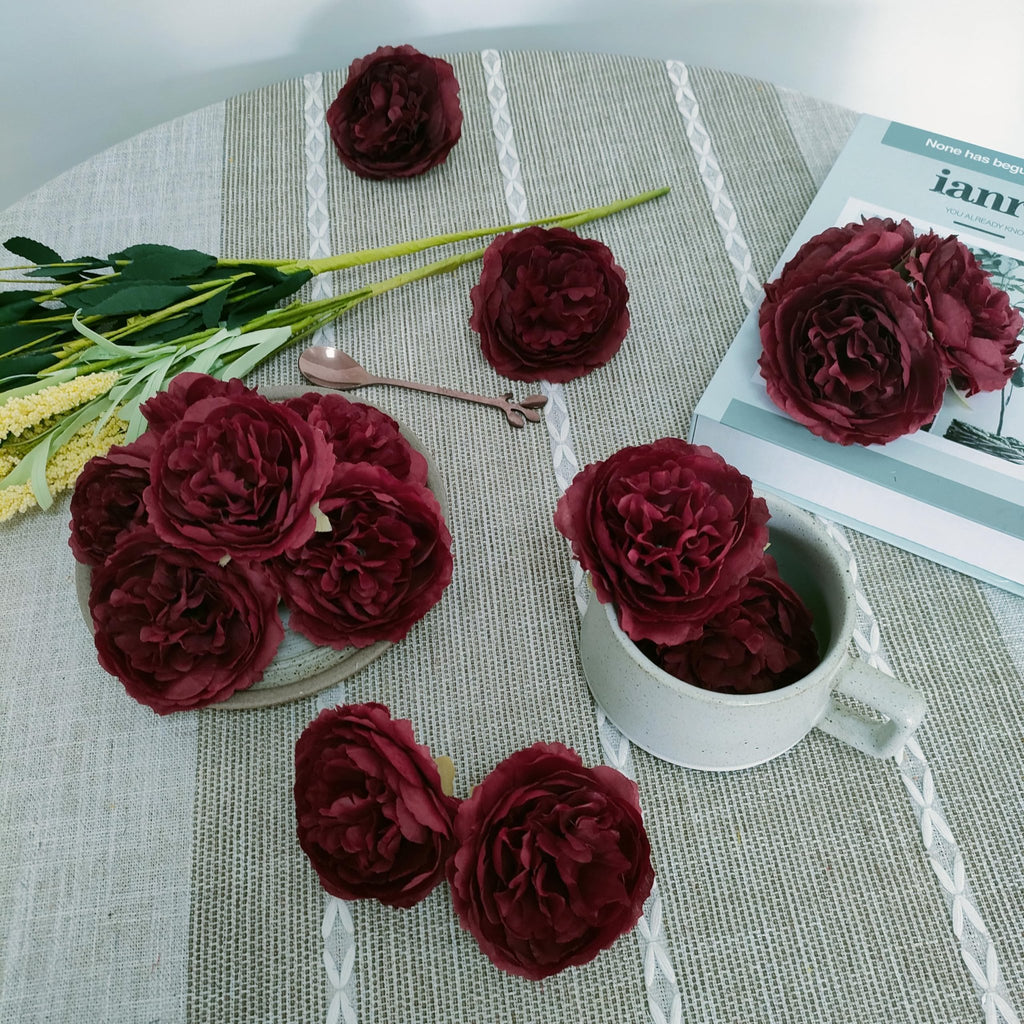 Burgundy artificial rose heads arranged on a textured tablecloth with a book and a mug.