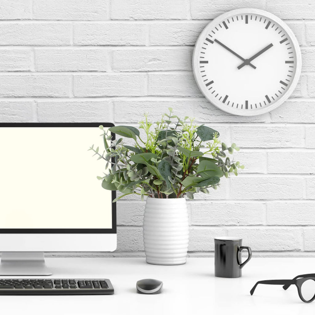 Artificial eucalyptus stems in a white vase on a desk with a computer, clock, and glasses