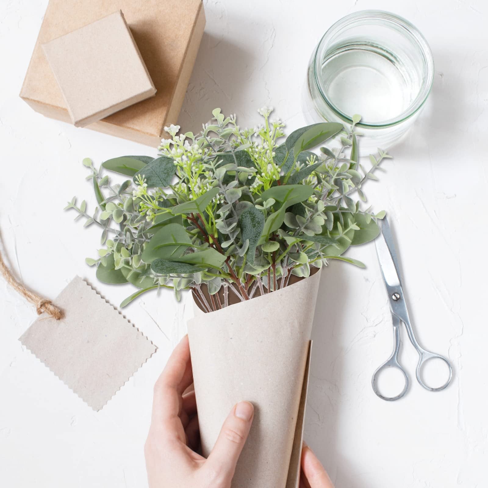 Hands holding a bouquet of artificial eucalyptus stems wrapped in brown paper, with gift boxes, a glass jar, and scissors on a white surface.