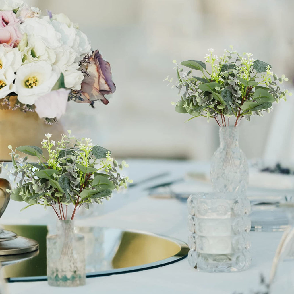 Two bouquets of artificial eucalyptus stems with silver dollar and baby leaves in clear vases on a table setting.