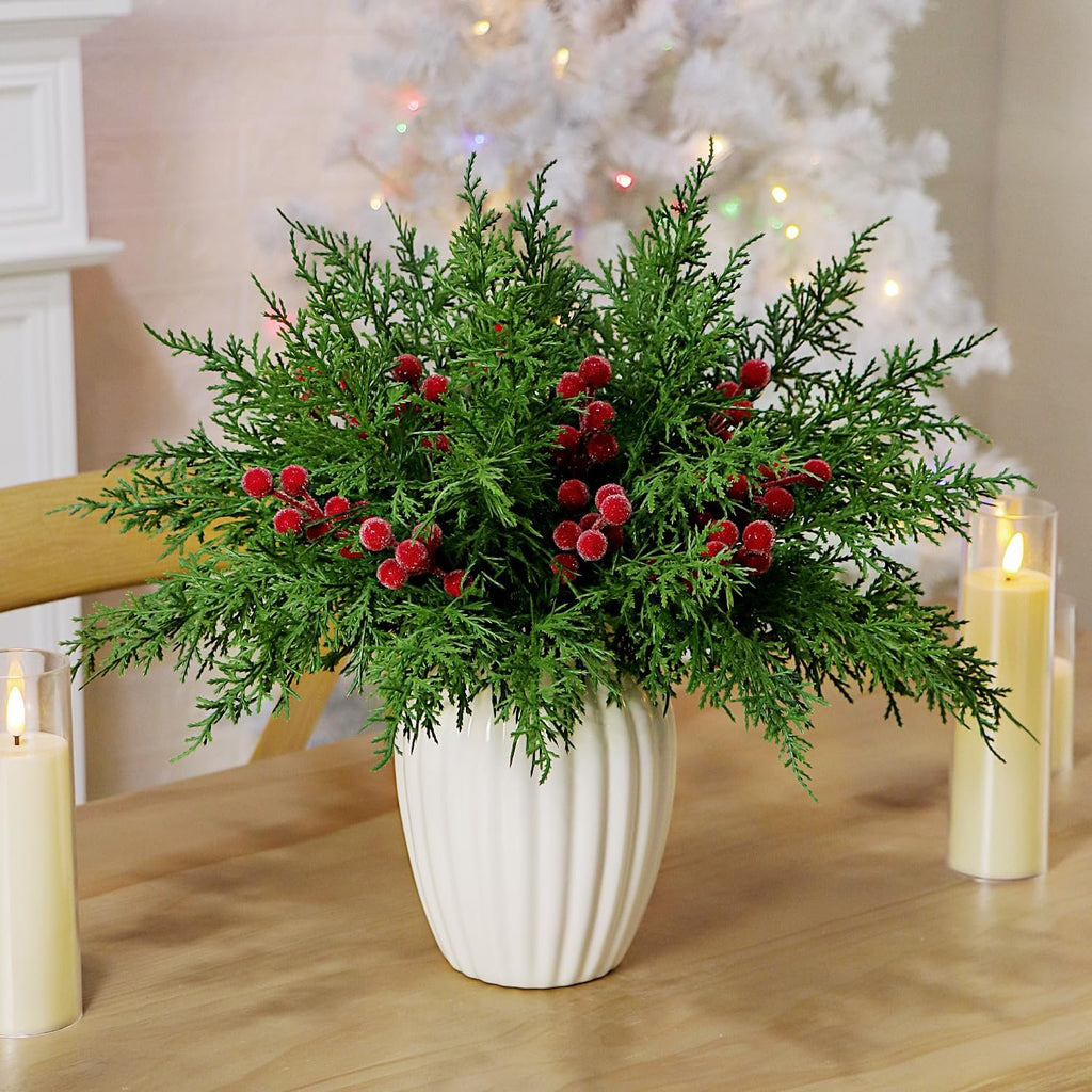 Artificial pine branches with red berries in a white vase on a wooden table with candles