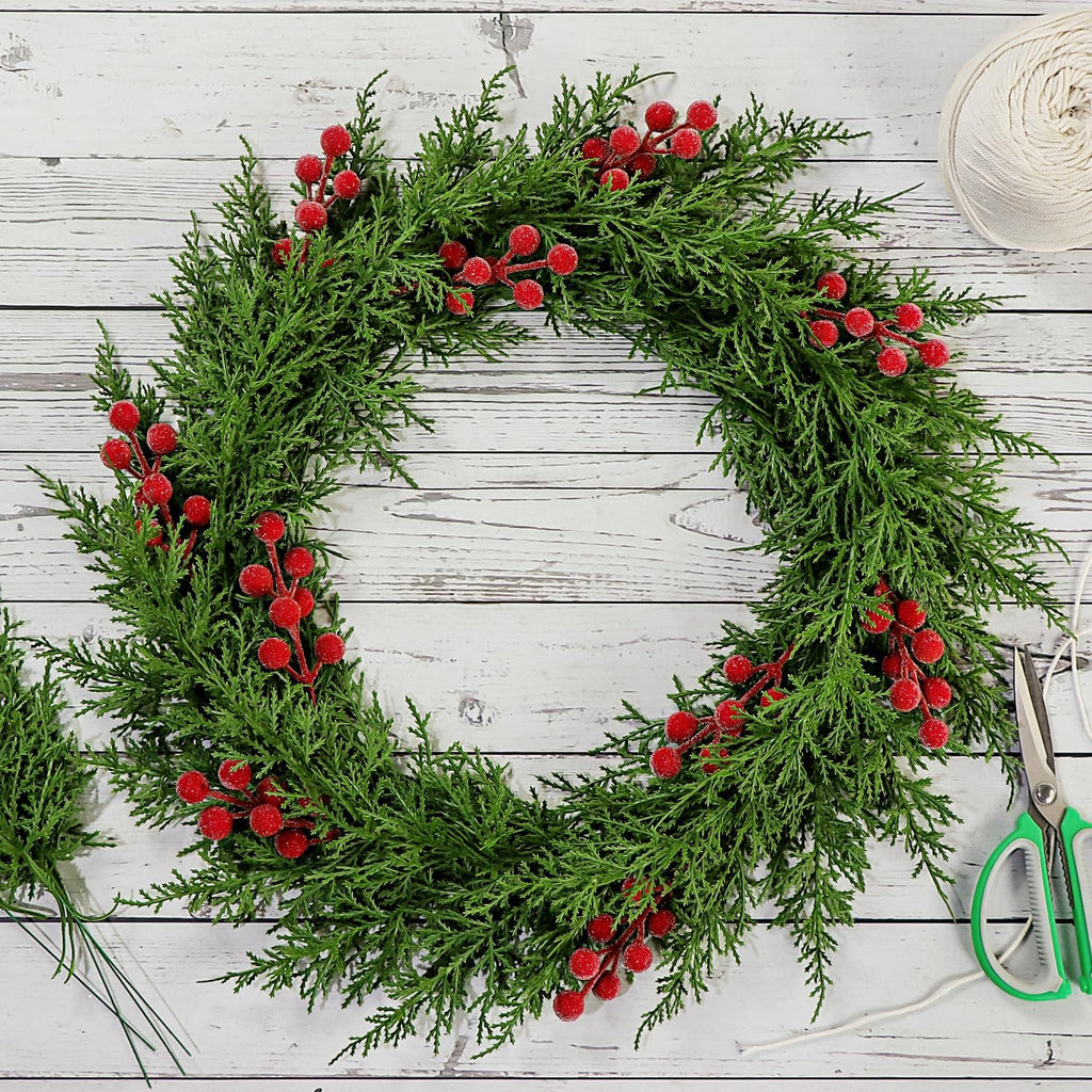 Artificial pine branch wreath with red berries, scissors, and a spool of twine on a white wooden background