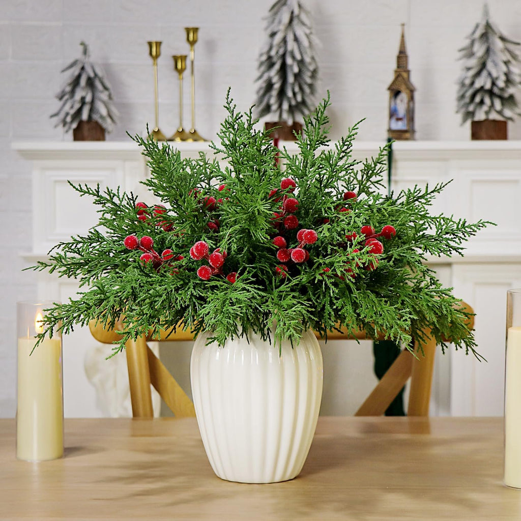 Faux cedar pine branches with red berries in a white vase on a wooden table with candles and Christmas decor