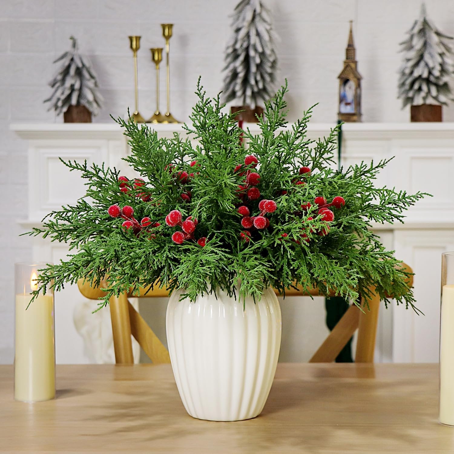 Faux cedar pine branches with red berries in a white vase on a wooden table with candles and Christmas decor