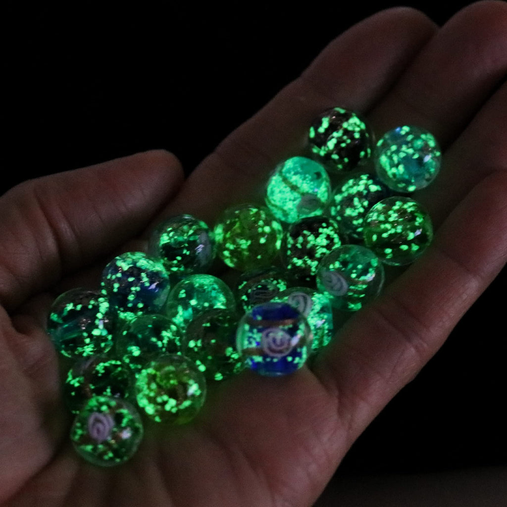 Hand holding a collection of glow-in-the-dark glass beads with green fluorescent sparkles and some with floral or blue patterns.
