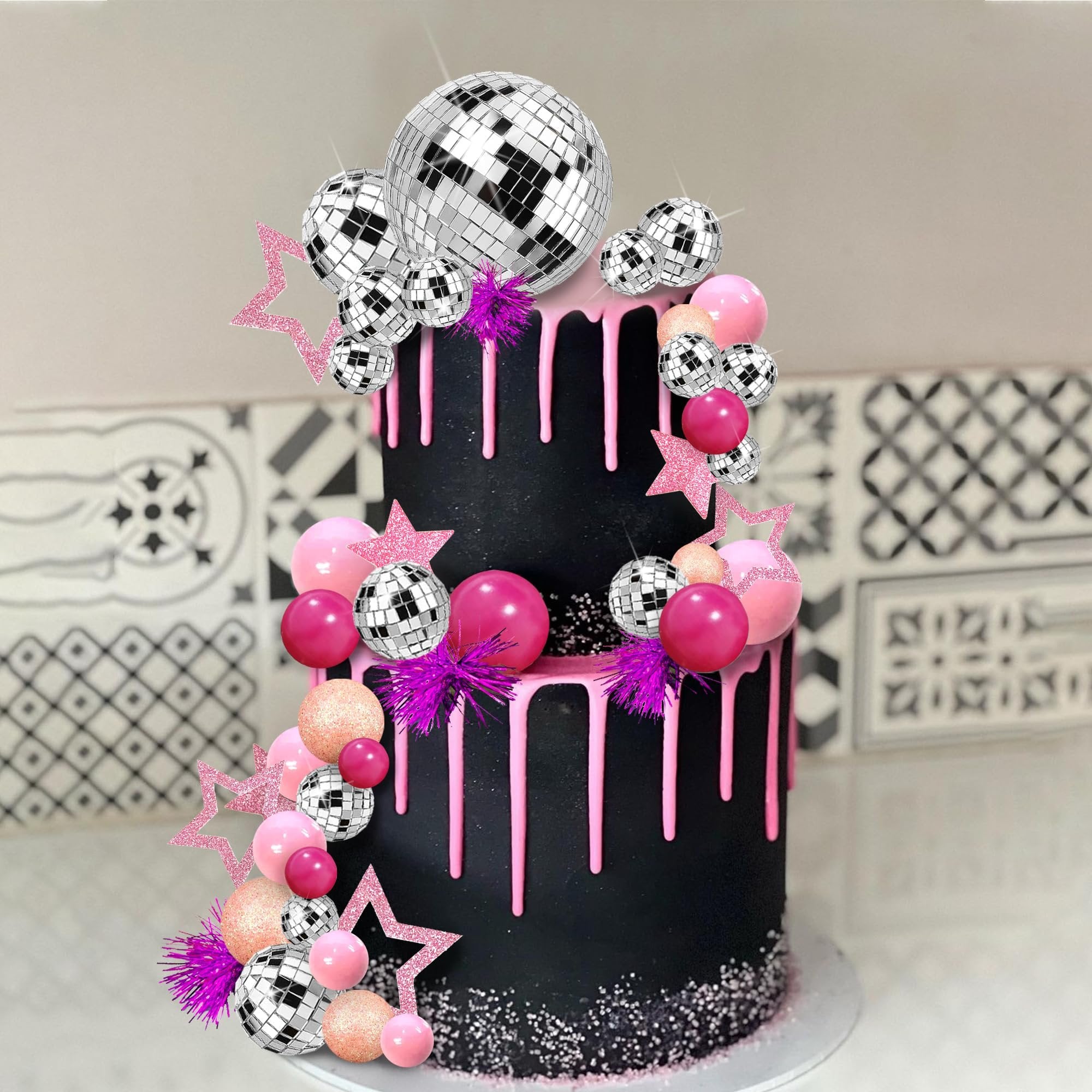 Two-tier black cake decorated with pink drip icing and a disco ball cake topper set featuring mirror balls, foam spheres, and star picks.