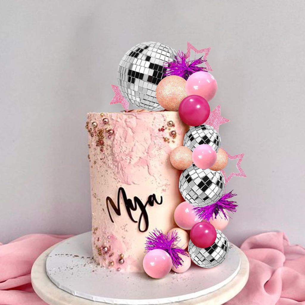 Pink disco ball cake topper set with mirror balls, foam spheres, glitter stars, and purple tinsel accents on a pink frosted cake with the name 'Mya' written in black script.
