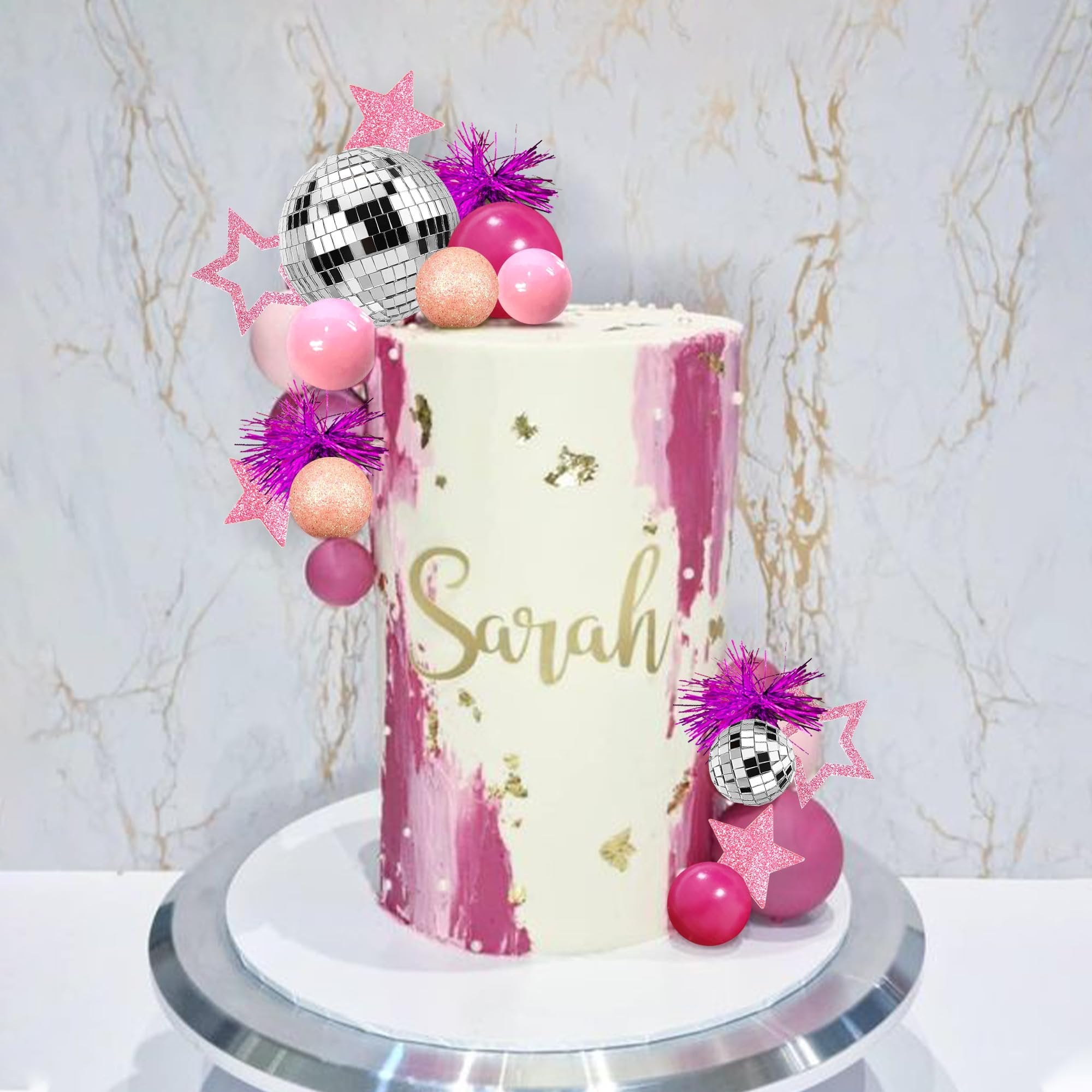 Tall cake decorated with pink frosting and gold flakes, topped with disco balls, pink spheres, and glitter stars.