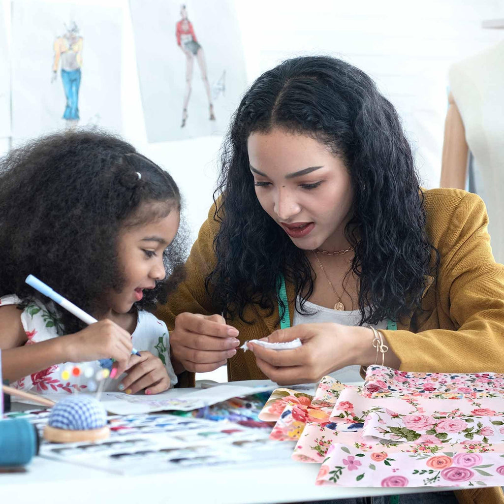 Woman and child crafting with pink floral fabric strips on a table with fashion sketches in the background.