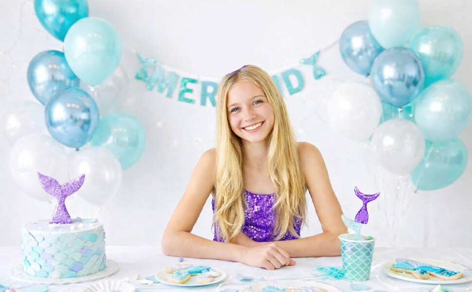 Smiling girl at a table with mermaid-themed birthday decorations including balloons, cake, and cookies.