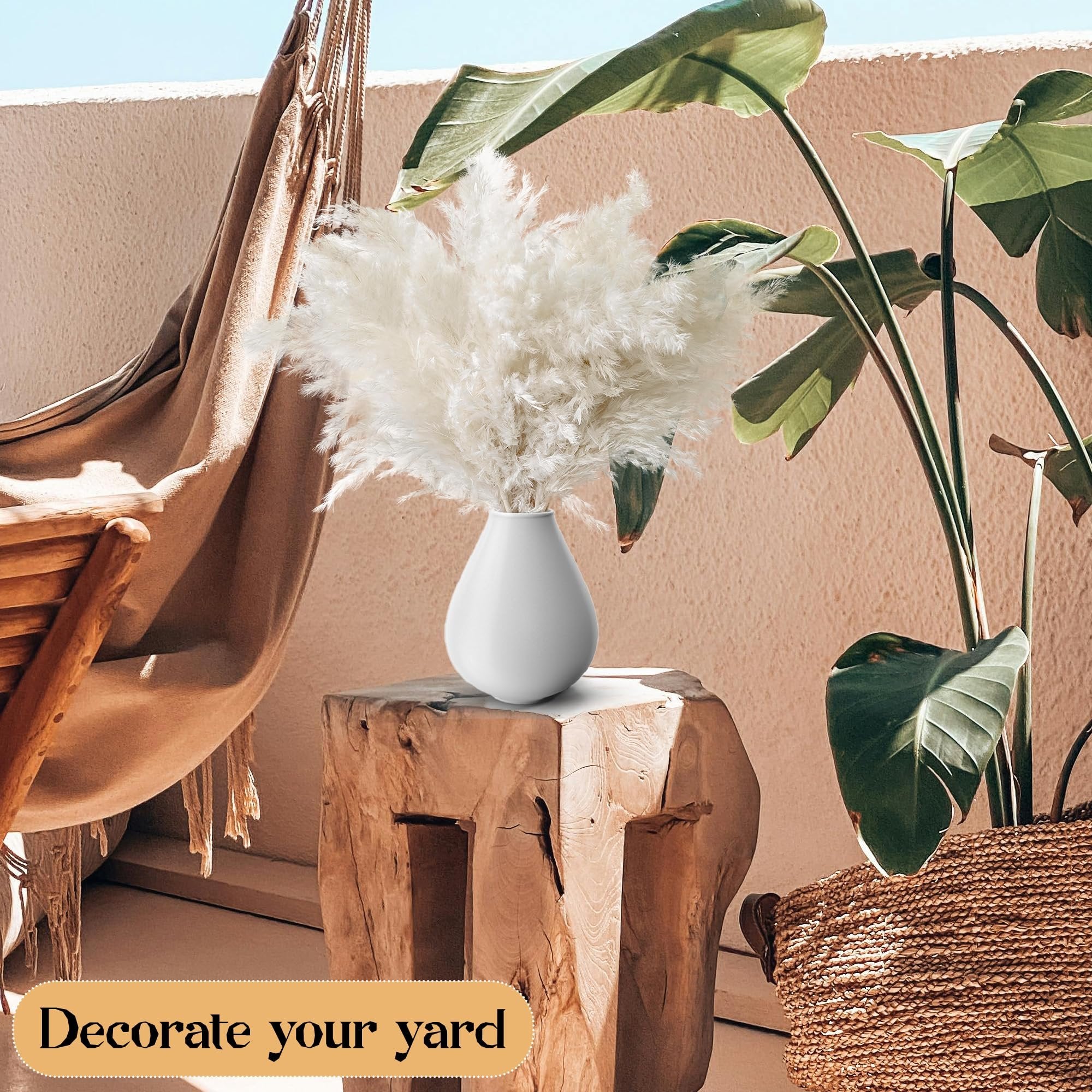 White pampas grass in a white vase on a wooden stool, with a hammock and large leaves in the background.