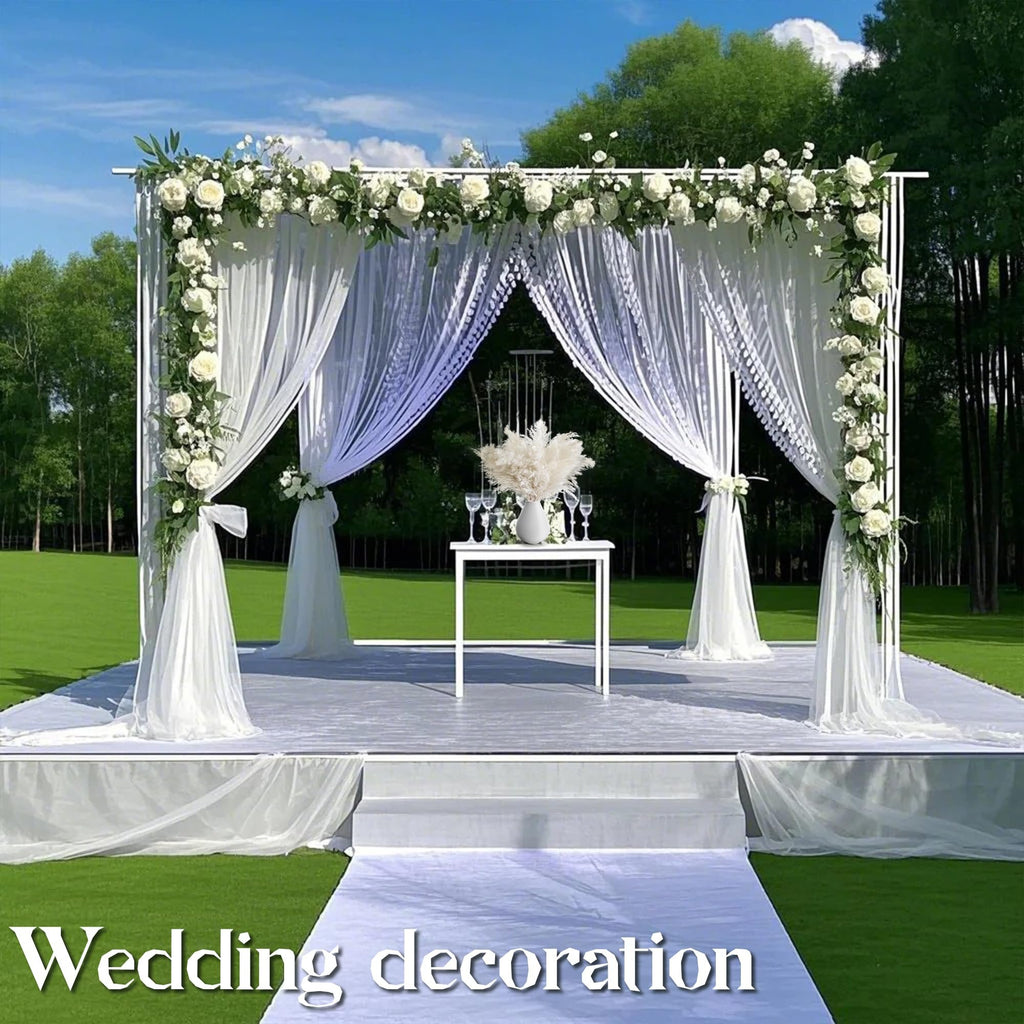 Outdoor wedding ceremony setup with white floral arch, draped fabric, and a table with pampas grass arrangement.
