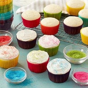 Decorated cupcakes in colorful paper liners with bowls of sprinkles, arranged on a surface.